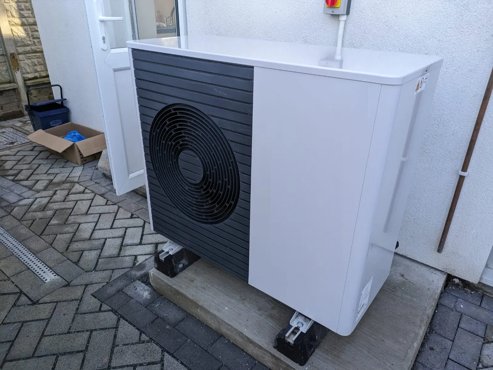 Photo of a white air-source heat pump outdoor unit with a dark fan grille, standing on a concrete pad beside a house wall, brand markings removed, shown for illustration