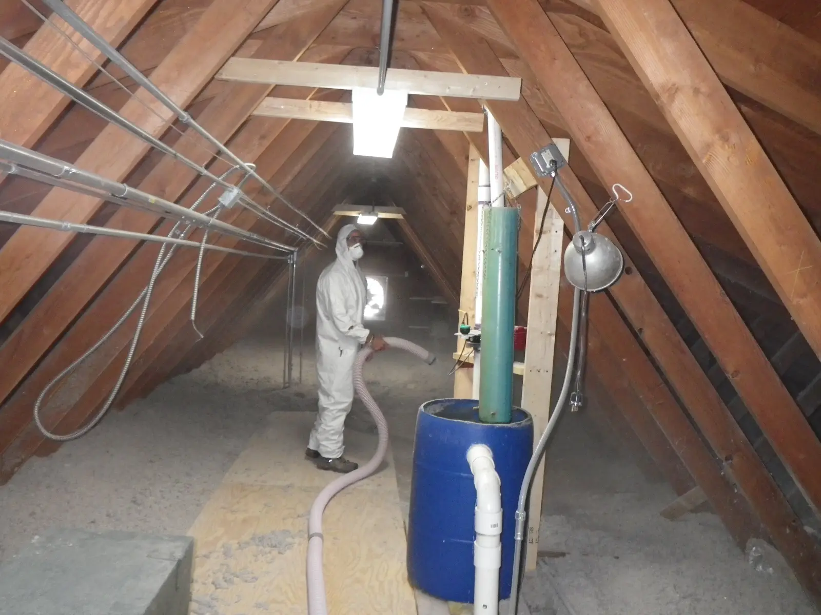 Photo of a worker in a white protective suit and mask blowing loose cellulose insulation across an attic floor through a long hose, shown for illustration