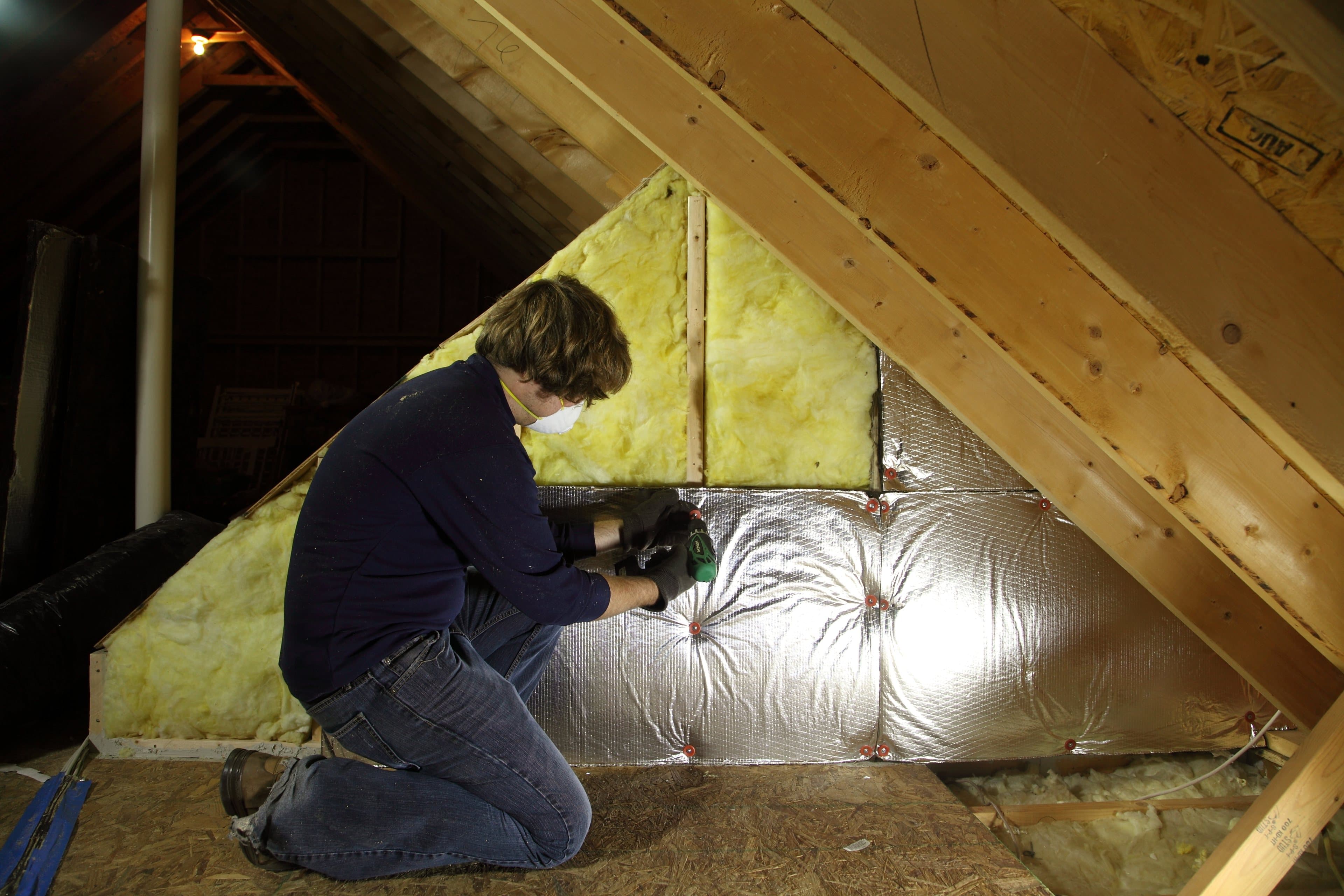 Photo of a worker in a dust mask fastening foil-faced foam board over fiberglass batts on an attic knee wall, shown for illustration