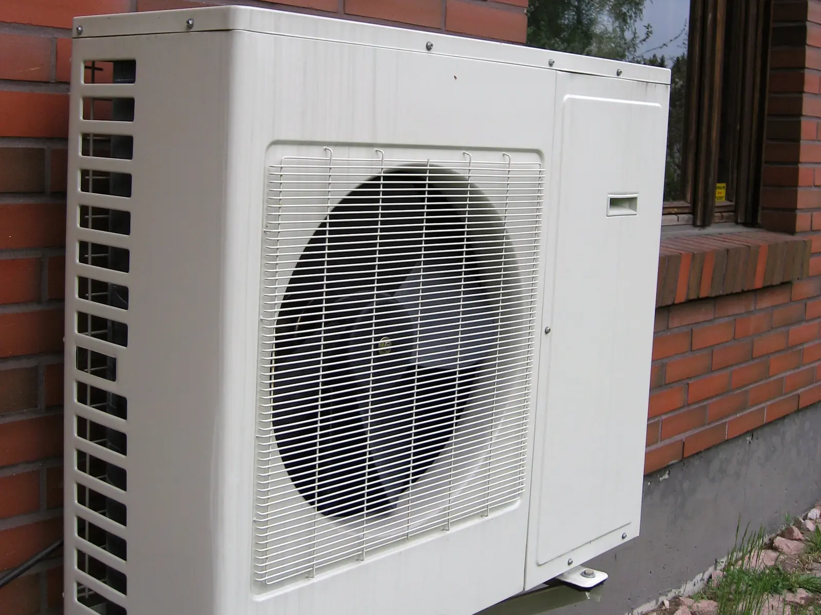 Photo of a ductless mini-split outdoor unit on a steel wall bracket against a brick house wall, shown for illustration