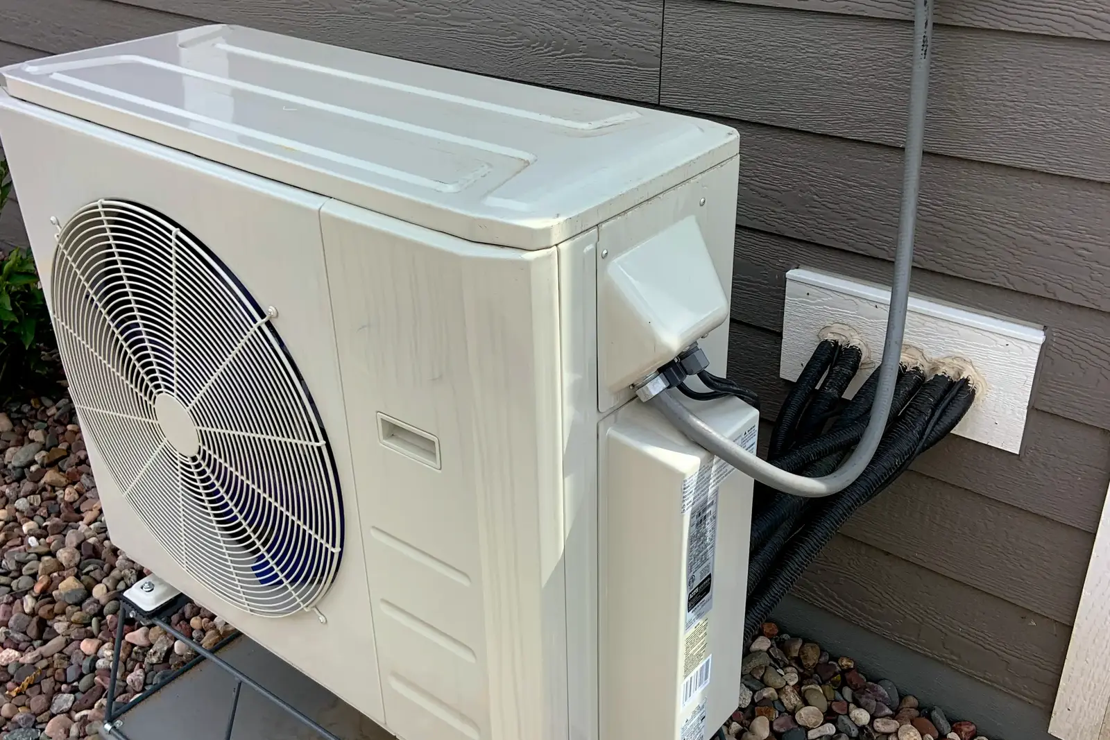 Photo of a ductless heat pump outdoor unit on a raised metal stand beside a house, with refrigerant lines running into the wall, shown for illustration