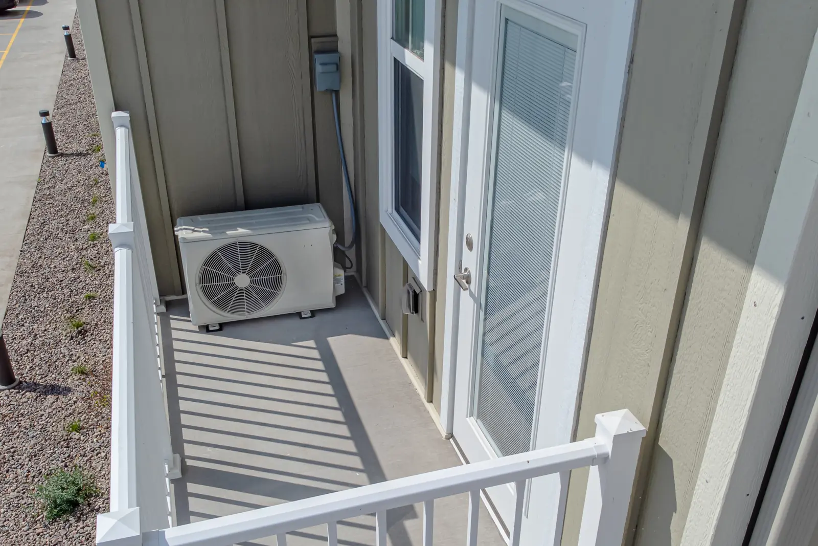 Photo of a small mini-split outdoor unit on a balcony beside a door, with its disconnect box on the wall above, shown for illustration