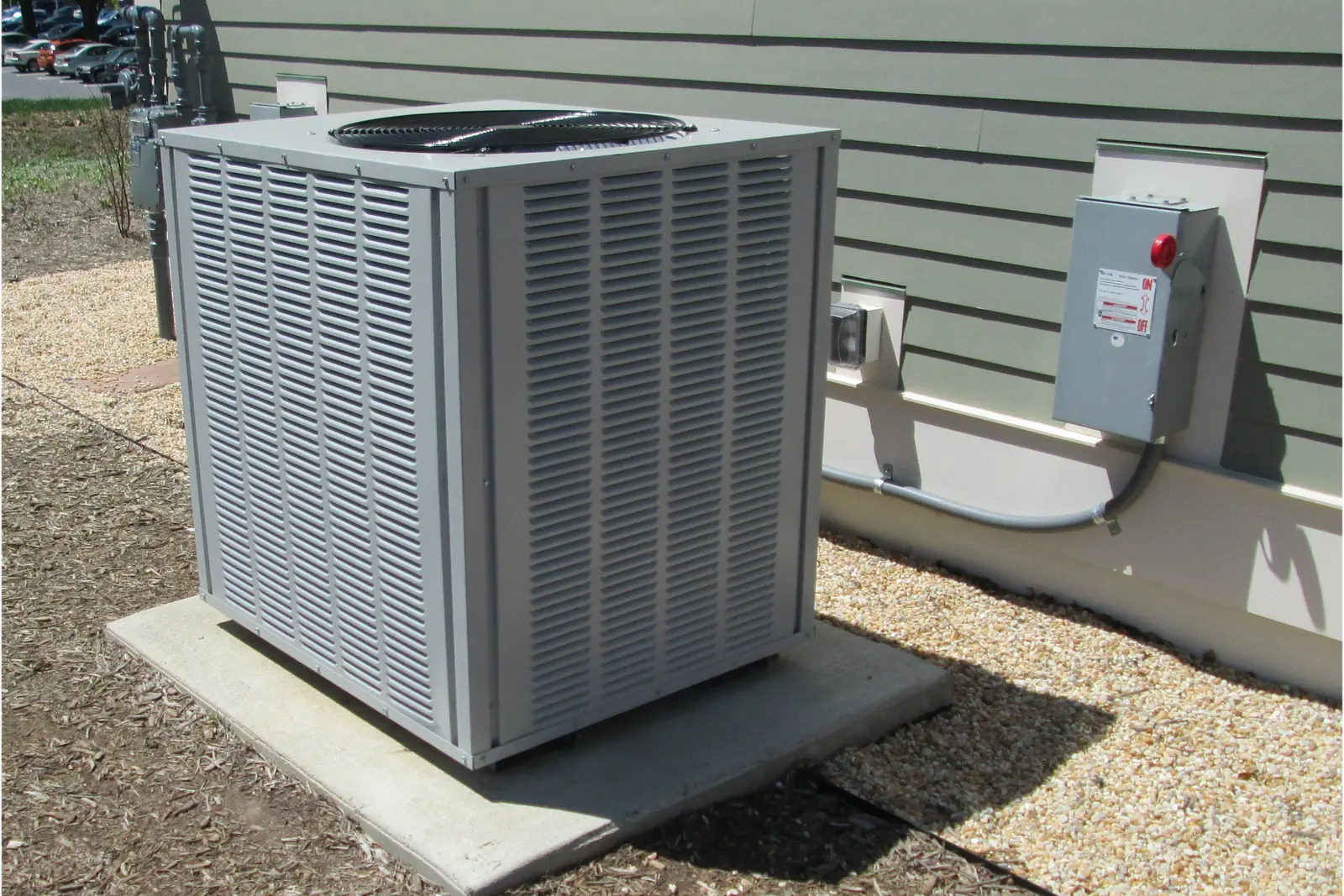 Photo of an outdoor heat pump or air conditioner unit on a concrete pad, with a wall-mounted electrical disconnect switch beside it, shown for illustration